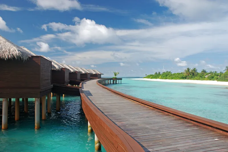 Holzsteg mit Wasserbungalows auf den Malediven führt über türkisblaues Wasser zu einer grünen Insel.