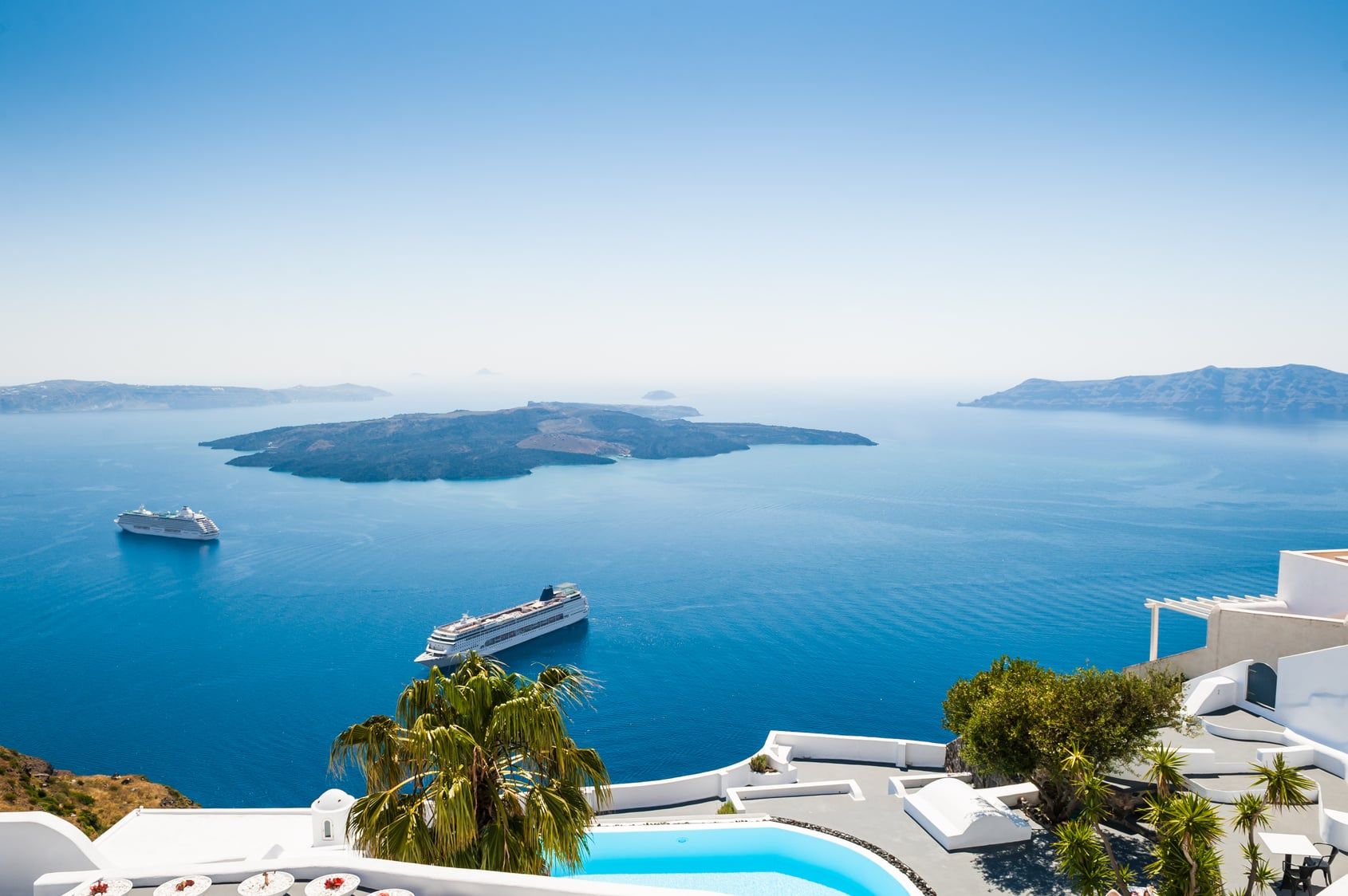 Blick von einer Terrasse auf Santorin mit Pool und zwei Kreuzfahrtschiffen in der Caldera.