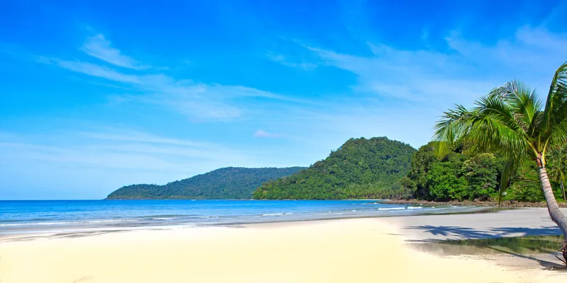 Weißer Sandstrand mit Kokospalme und grünem Bergvorsprung an einer ruhigen tropischen Bucht in Südostasien.